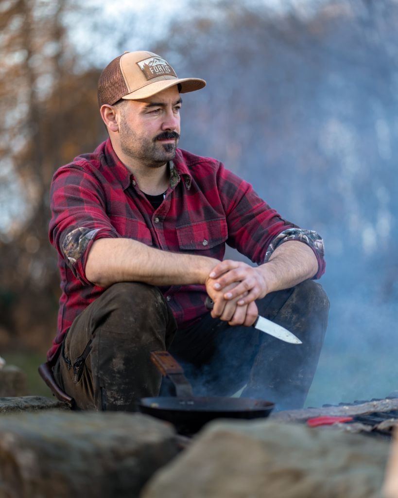 Cai Ap Bryn sitting by a campfire holding a knife
