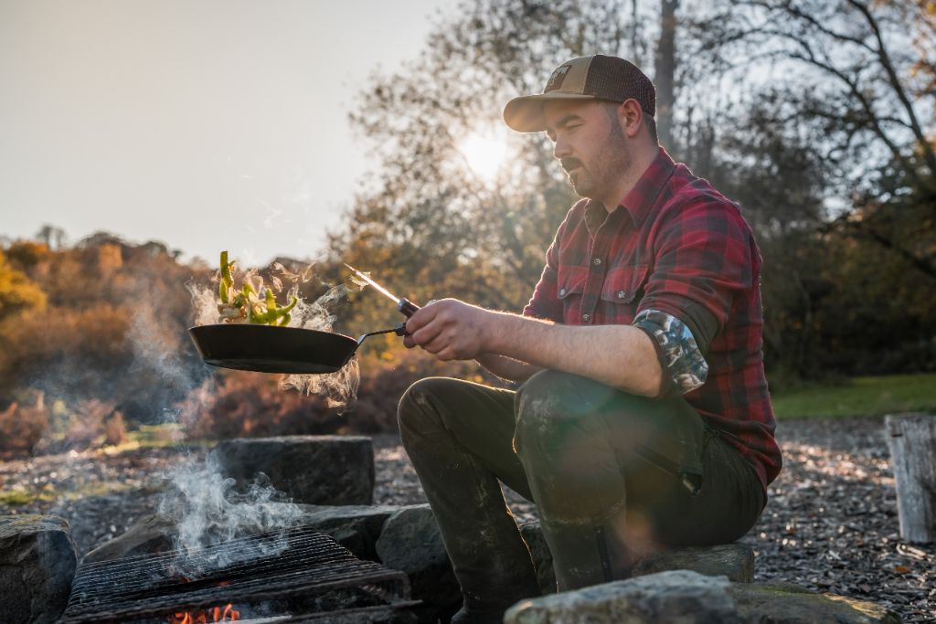 Cai Ap Bryn tossing food in a pan over an open campfire at sunset