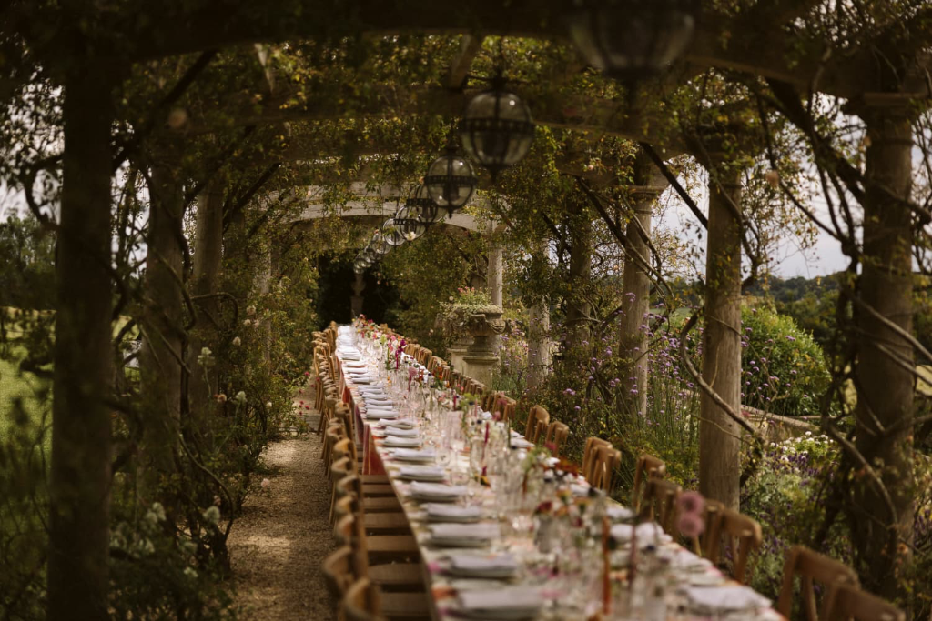 Long wedding banquet table under a vine-covered pergola with lanterns and wildflowers